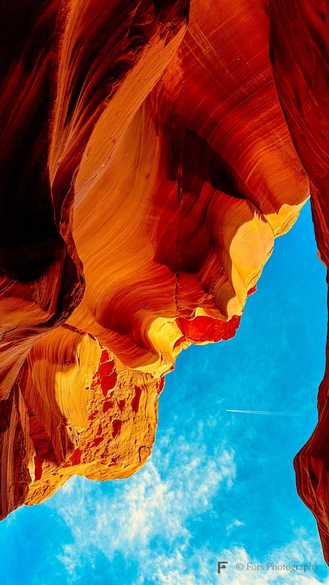 Looking straight up inside a slot canyon: curving orange and gold sandstone walls frame a patch of bright blue sky with white clouds and a thin jet trail.
