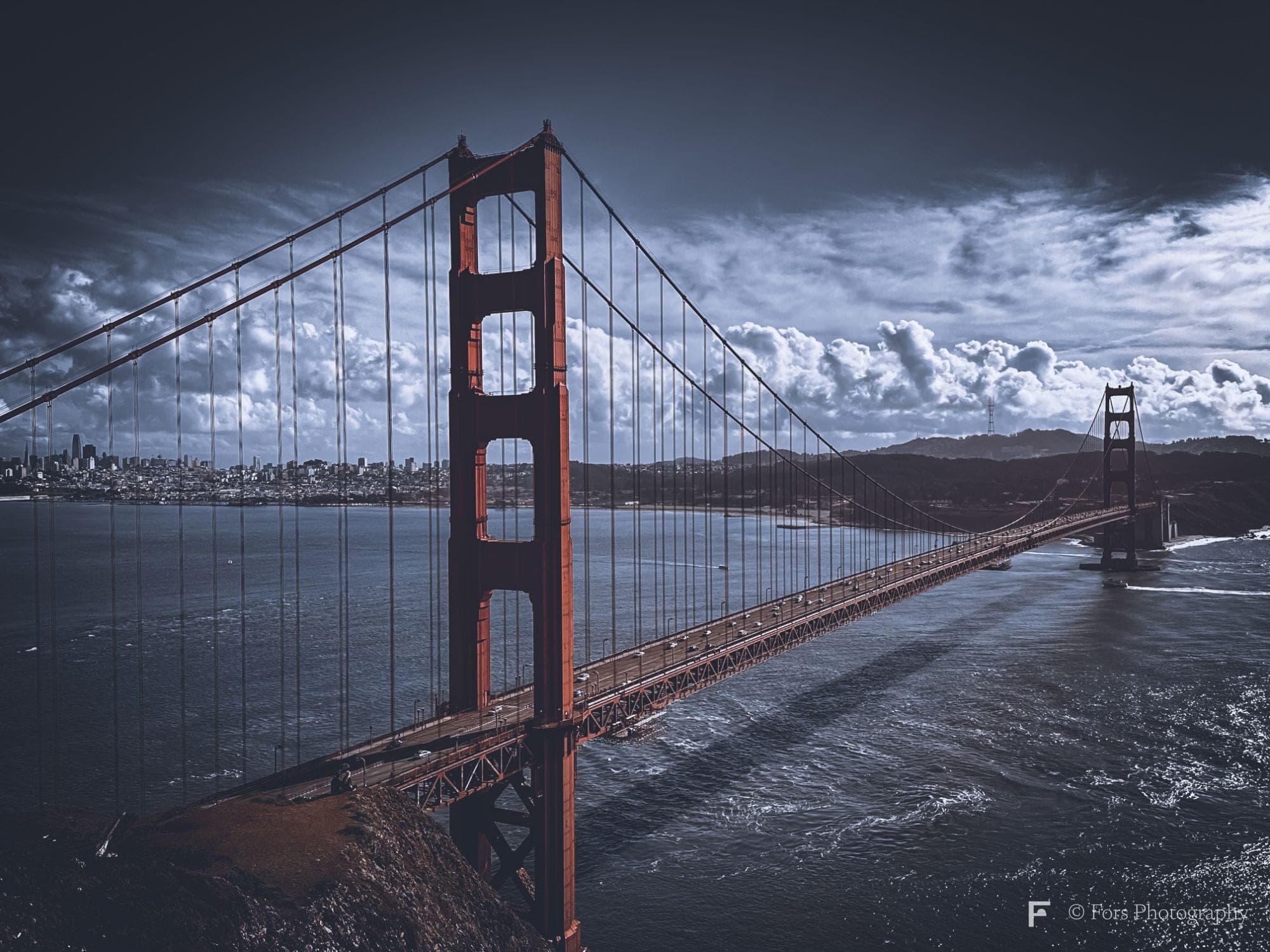 The Golden Gate Bridge seen from above its north tower, the full span crossing dark water toward the San Francisco skyline under heavy clouds.