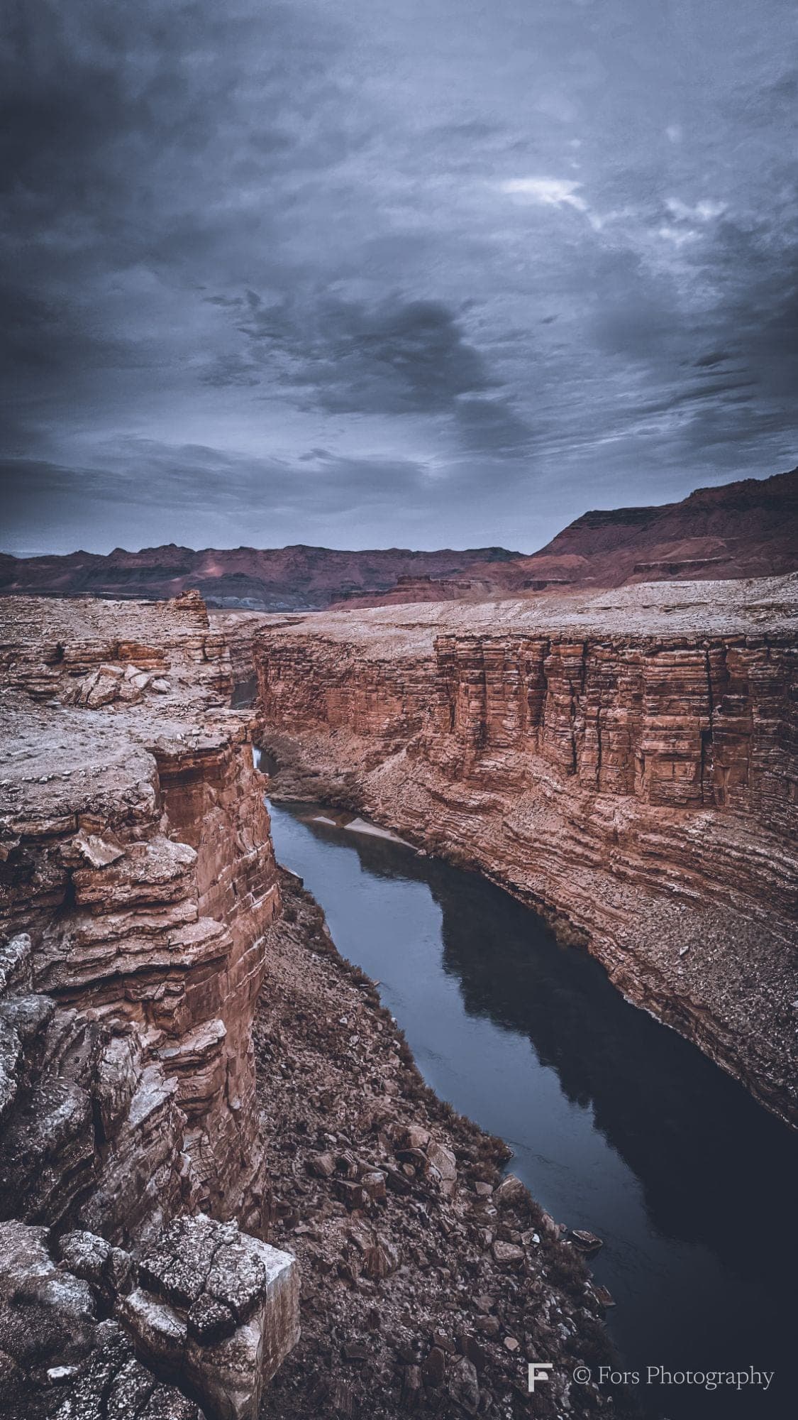 A dark river winding between layered red sandstone cliffs under a stormy blue-grey sky, with distant mesas on the horizon.