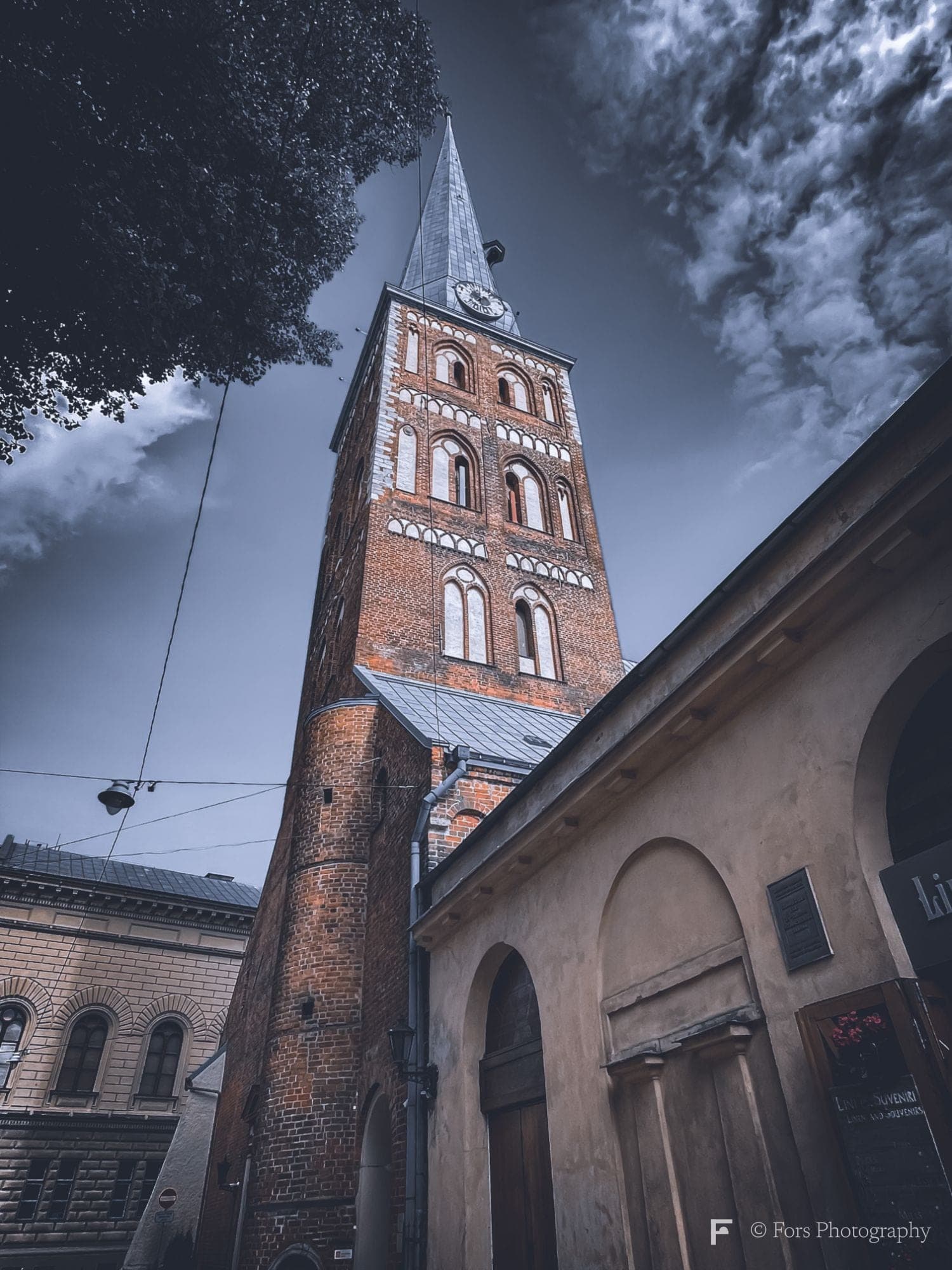 A tall red-brick church tower with a pointed spire and a clock, seen from below between a tree and a pale arched building, against a dark blue sky with heavy clouds.